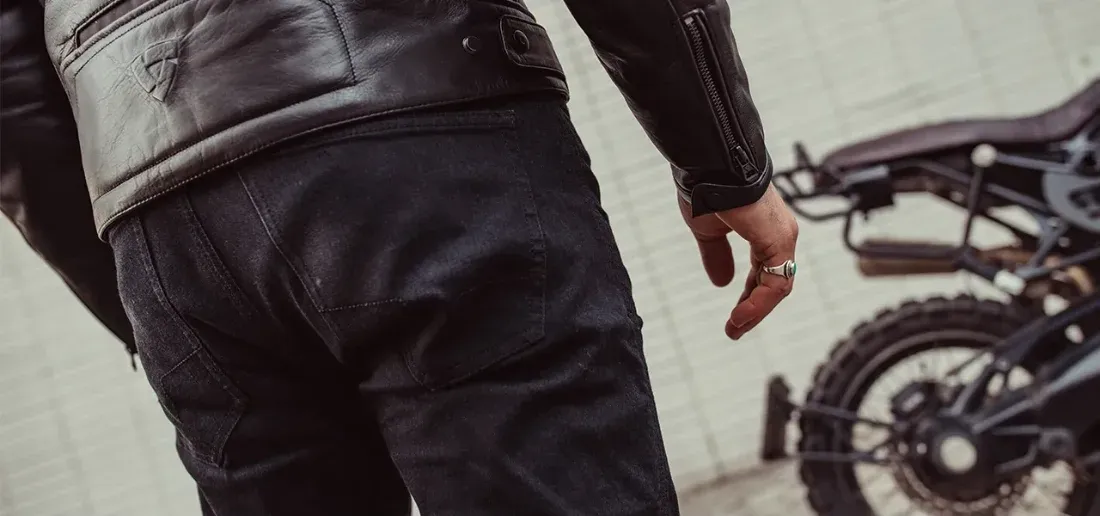 Detaliu real cu blugi REVIT Jackson 2 SK purtați lângă motocicletă, cu fundal real și zona șoldului în prim-plan